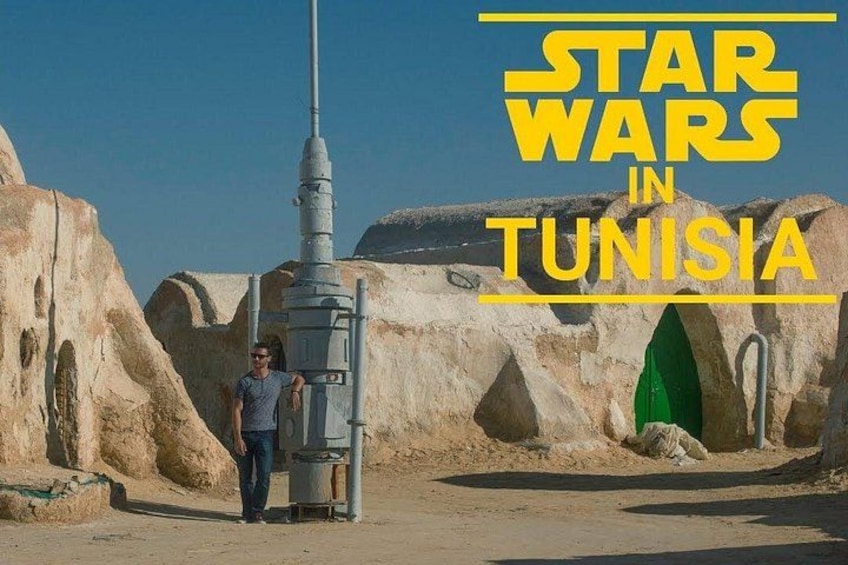 A desert landscape with a large structure resembling a Star Wars Tatooine-style cantina, a person standing next to it, and a green door in the background. 3