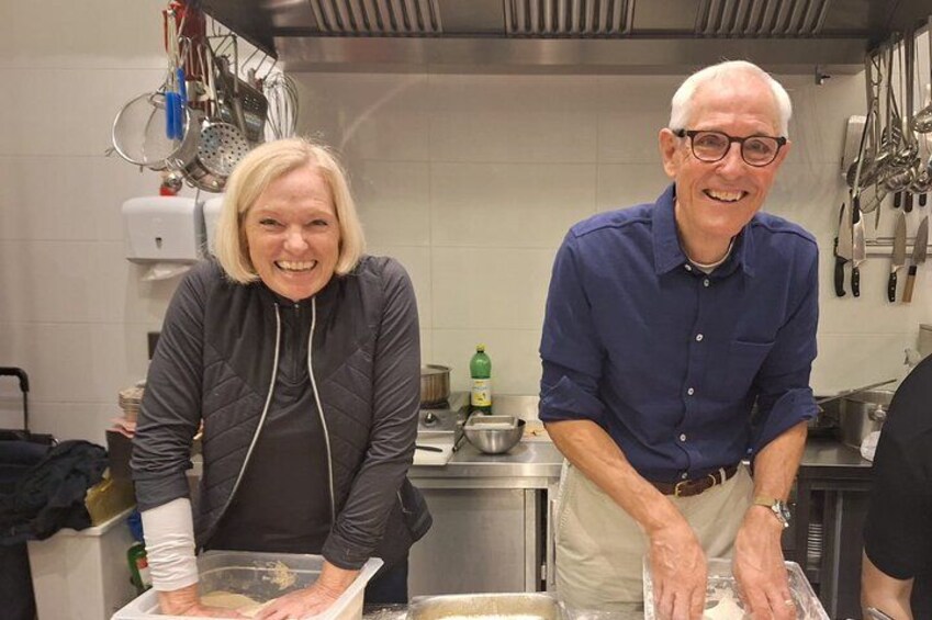 Dos personas en una cocina, una vestida con una chaqueta oscura y la otra con una camisa azul, con diversos utensilios y electrodomésticos de cocina al fondo. 4