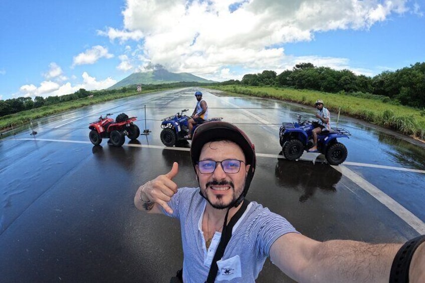A person giving a thumbs-up gesture, two quad bikes on a wet road, and a mountain in the background. 1