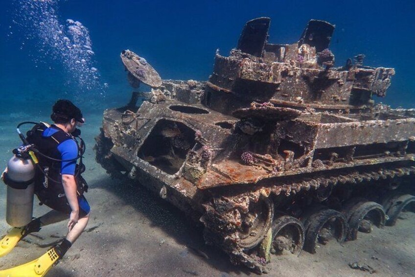 A diver with a scuba tank and fins near a sunken military tank underwater. 4