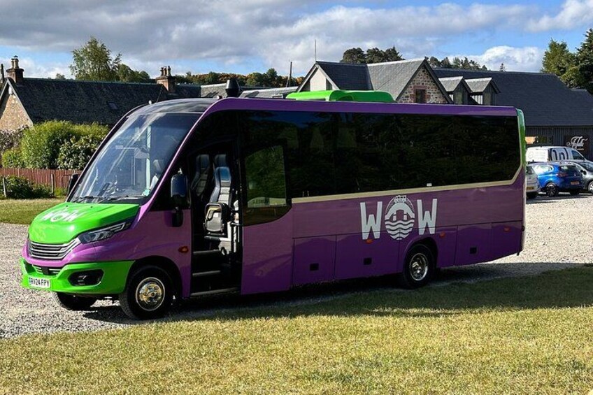 Un autobus aux couleurs violettes et vertes est garé sur une pelouse, avec un bâtiment en arrière-plan. 3