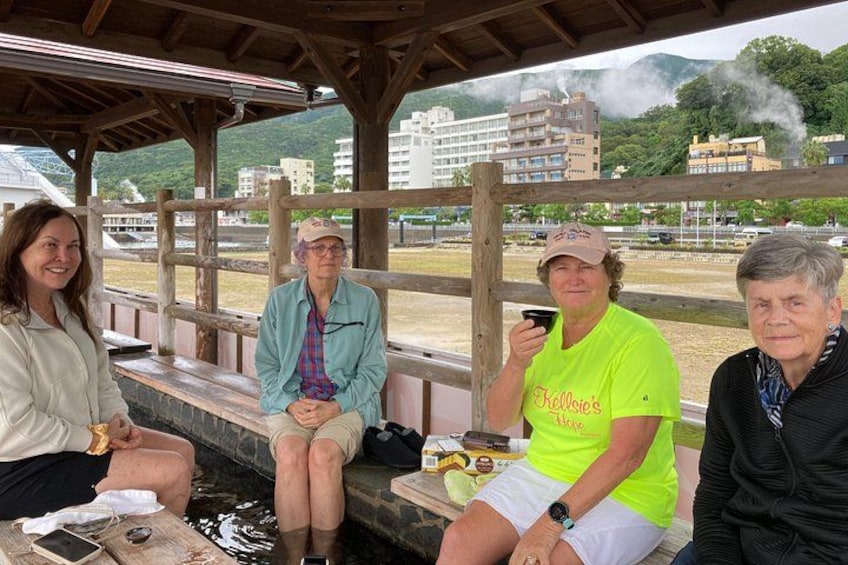 Three people sitting on a wooden bench at a hot spring with steam rising in the background. 3