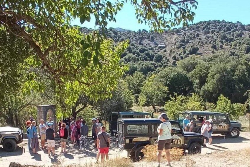 A group of people standing near a vehicle, a stone structure, and trees in a hilly area. 4
