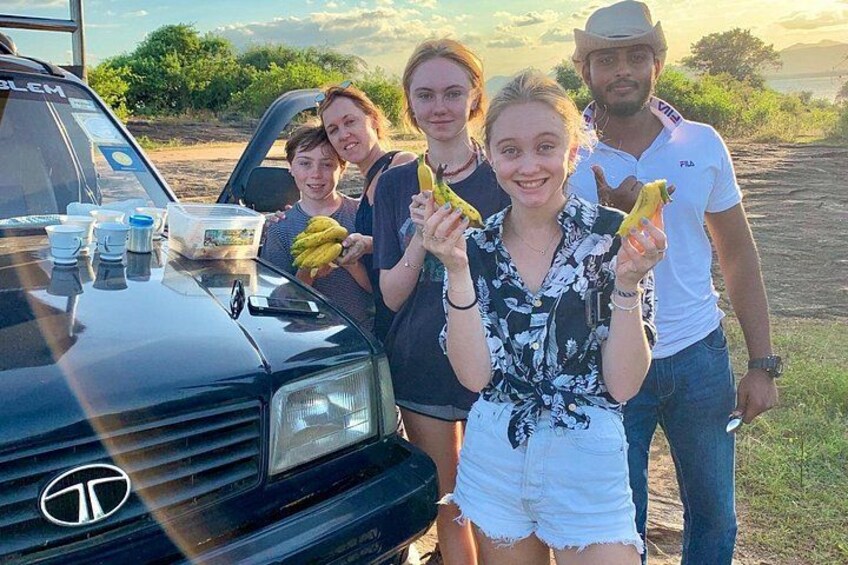 Un groupe de personnes se tient à côté d’un véhicule sur le capot duquel on peut voir des bananes et une bouteille. 1