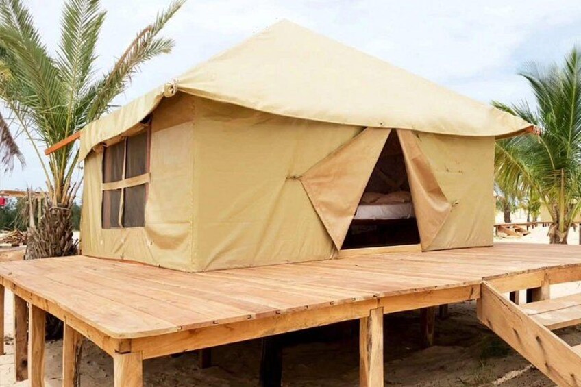 Une tente de couleur beige avec une terrasse en bois, des palmiers et, au fond, une plage de sable. 3