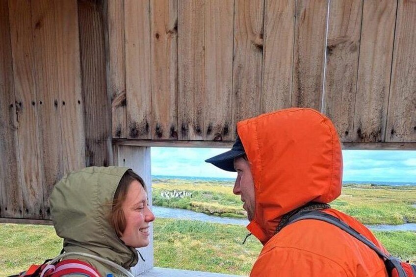 Two people in outdoor clothing, one in a green jacket and the other in an orange jacket, standing in front of a wooden structure with a view of a grassy landscape and a body of water. 5