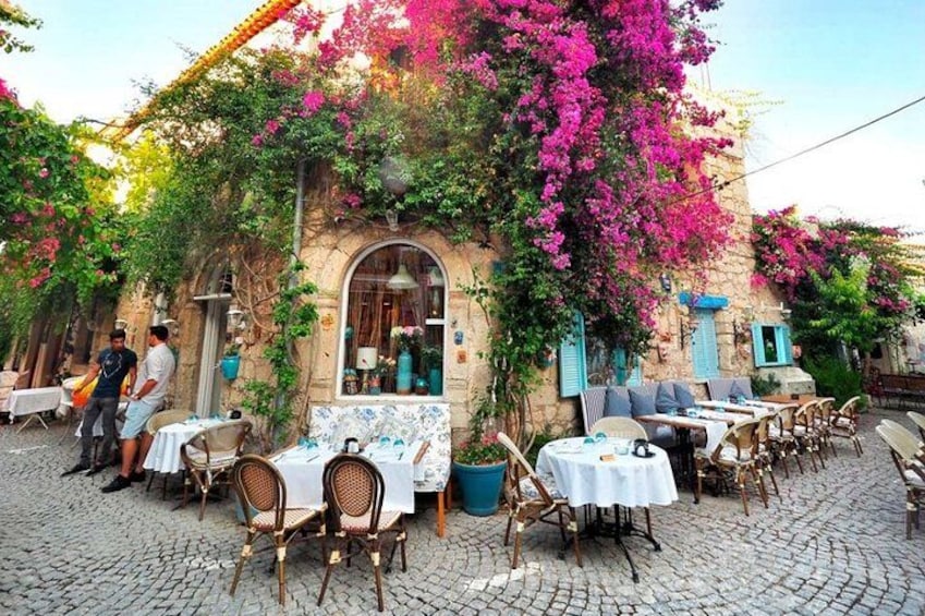 An outdoor café with tables and chairs, a stone building with a window, and vibrant pink bougainvillea flowers. 5