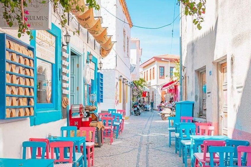 A narrow street with colorful chairs and tables, a blue building with a sign, and a motorbike in the distance. 1