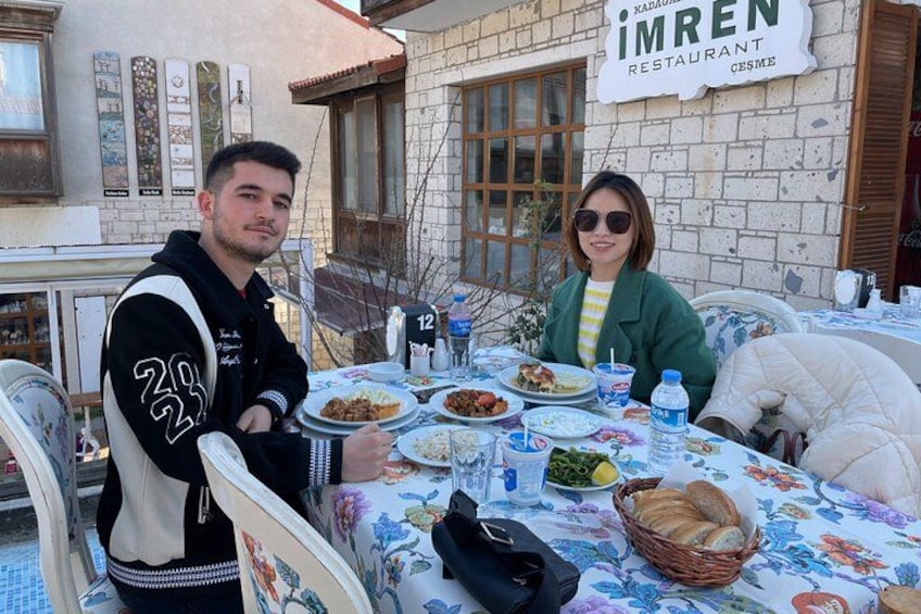 Two people dining at a table with a floral tablecloth, plates of food, and a sign for 'IMREN RESTAURANT' in the background. 2