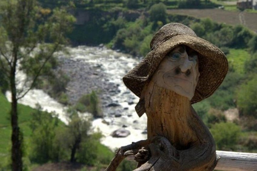 A wooden sculpture of a bird wearing a hat, overlooking a river with a forested landscape in the background. 2