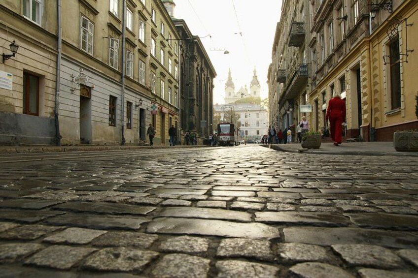 A cobblestone street with buildings on either side, a person in red, and a distant church spire. 5