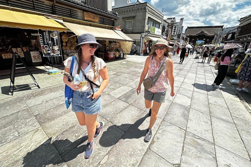 Two people walking on a paved street with shops and buildings in the background. 1