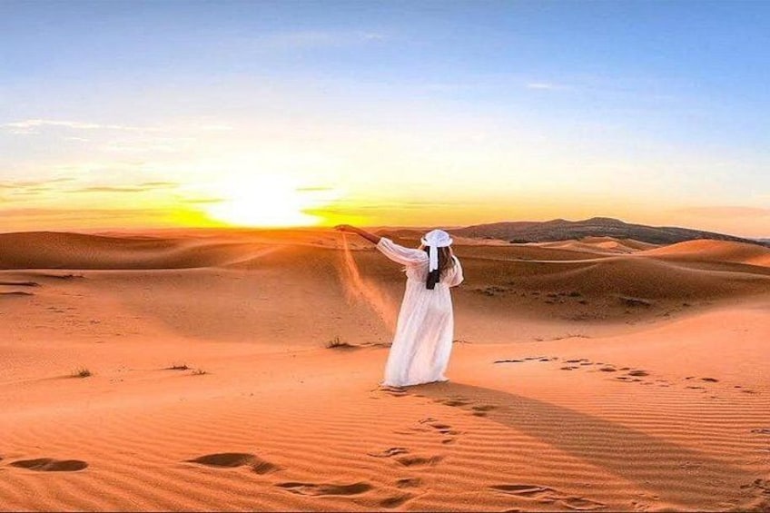 A person in a white dress standing in a desert with sand dunes and a sunset in the background. 5