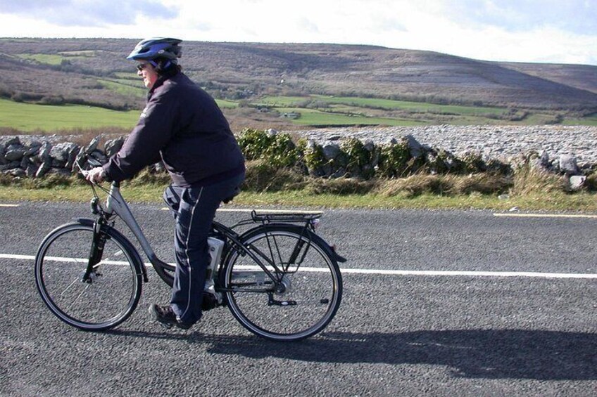 A person riding a bicycle on a paved road with a stone wall and grassy fields in the background. 1