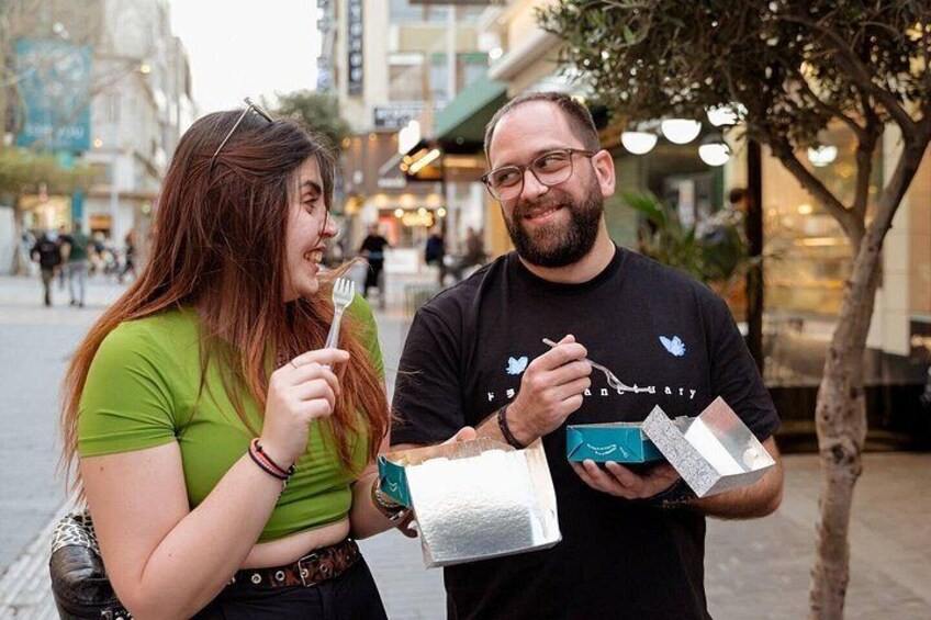 Two people holding shopping bags in an urban setting with buildings and a tree in the background. 2