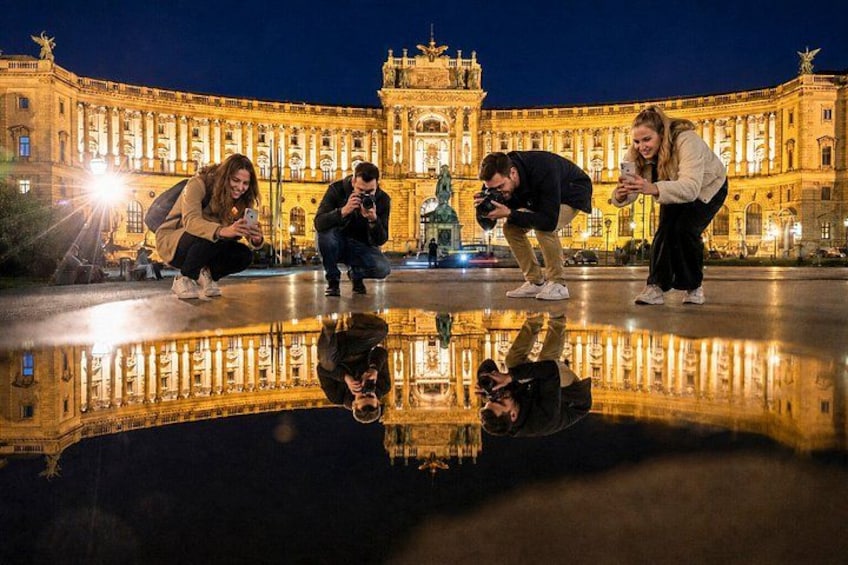 A historic building with a reflection in a puddle, people taking photos, and a statue in the foreground. 1