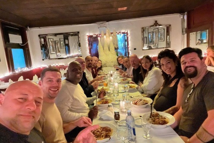 A group of people seated around a long dining table in a restaurant with a white tablecloth, plates of food, and glasses of water. 4