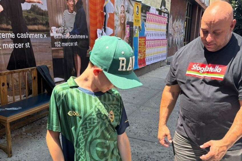 A person wearing a green sports jersey and a cap with 'BRA' on it, standing on a sidewalk. 2