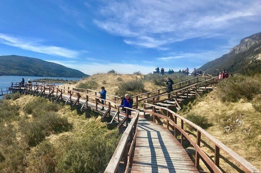 A wooden boardwalk with people walking and sitting, overlooking a lake and hills. 1