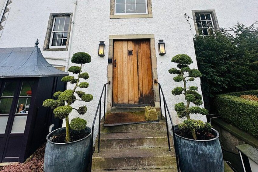 A white building with a wooden door and steps leading up to it. There are two potted topiary trees on either side of the steps. 4