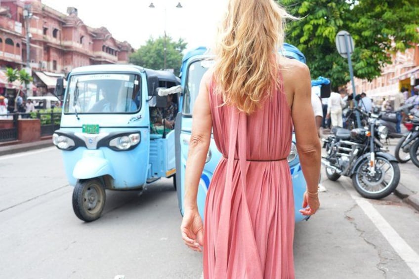 A woman in a pink dress walking on a street with a blue auto-rickshaw and motorcycles parked along the side. 3