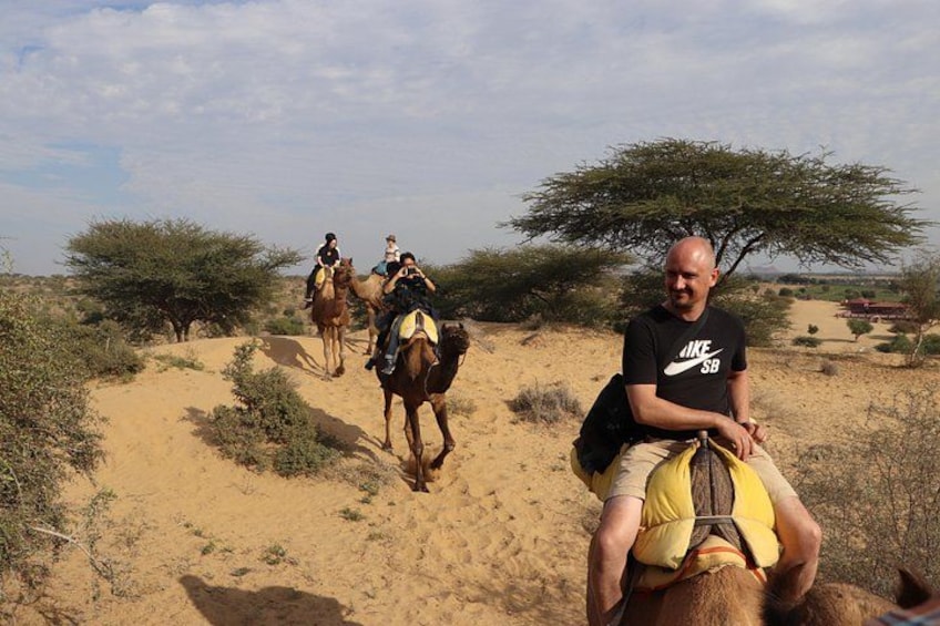 A man riding a camel in a desert with sparse vegetation and a clear sky. 1