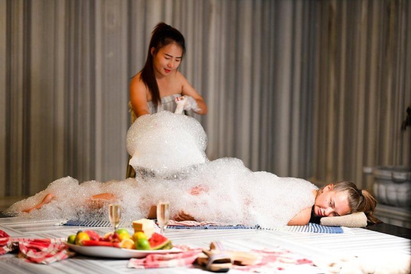 A person lying on a bed covered in bubbles, with a plate of fruit and a bottle nearby. 2
