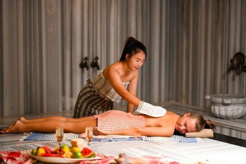 A person receiving a massage on a bed with a striped blanket, a bowl of fruit, and a stone basin in the background. 4