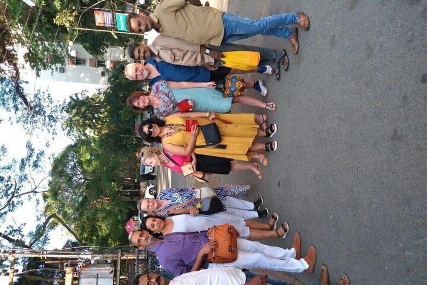 A group of people standing on a street, some holding bags, with trees and buildings in the background. 3