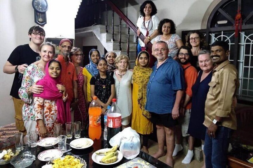 A group of people gathered around a table with food and drinks, in a room with a staircase and a clock on the wall. 5
