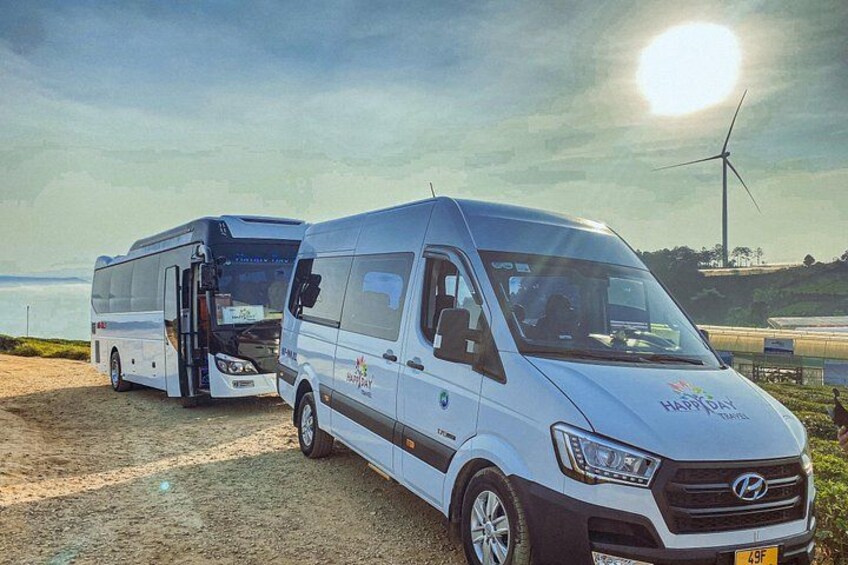 Two buses parked on a dirt road with a body of water and wind turbines in the background. 2