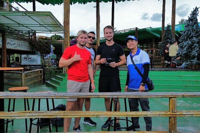 A group of people standing on a wooden platform with a green floor, a metal railing, and a building with a green roof in the background. 3