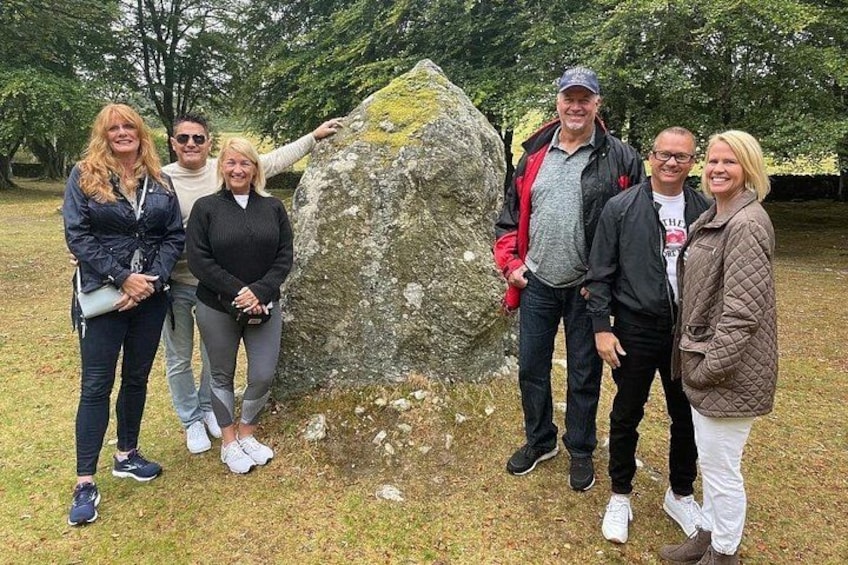 Een groep mensen staat voor een groot stenen monument in een grasrijk gebied, met bomen op de achtergrond. 5