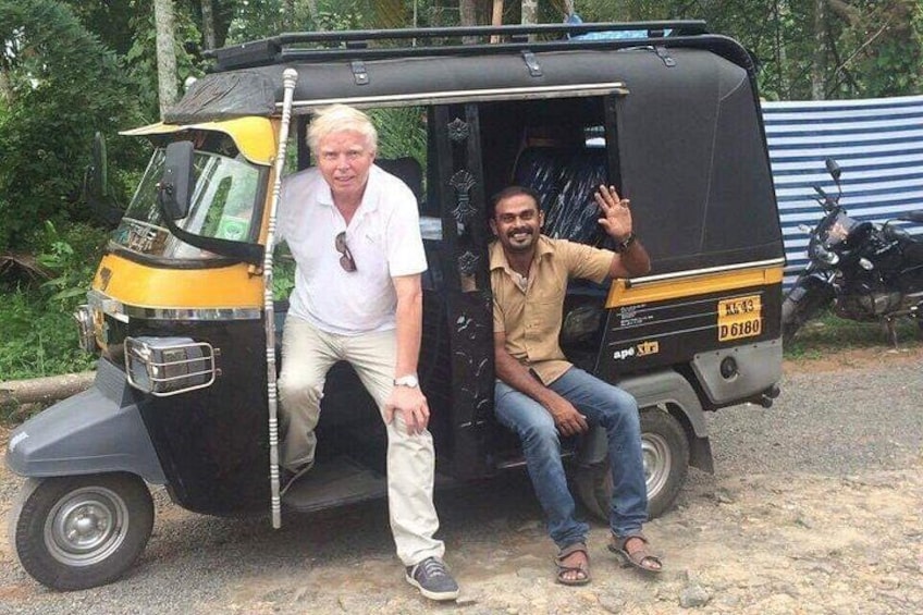 Two people in an auto-rickshaw, one standing and one sitting, with a motorcycle in the background. 1