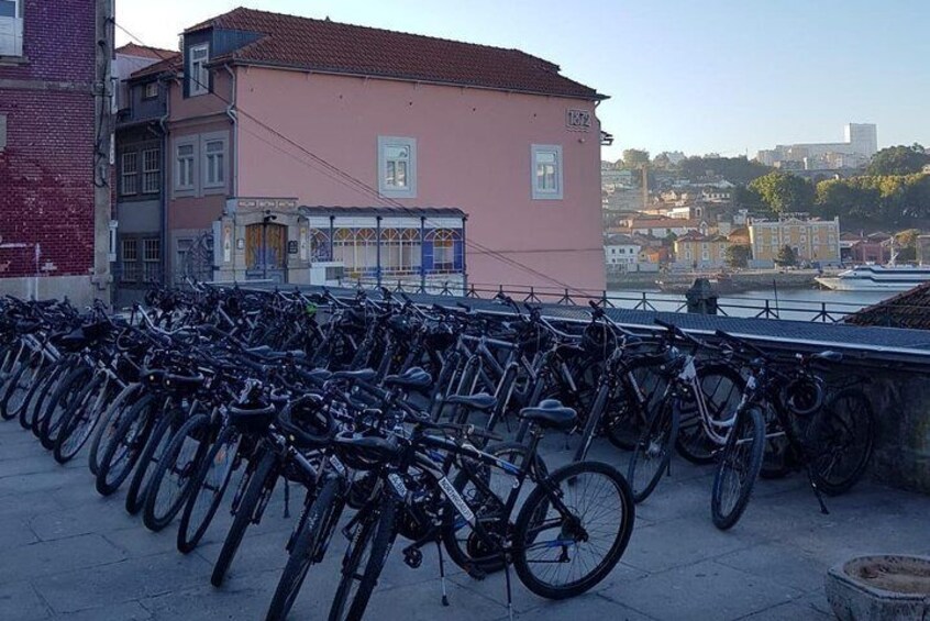 A row of bicycles parked on a sidewalk in front of a pink building with a view of a river and buildings in the background. 2