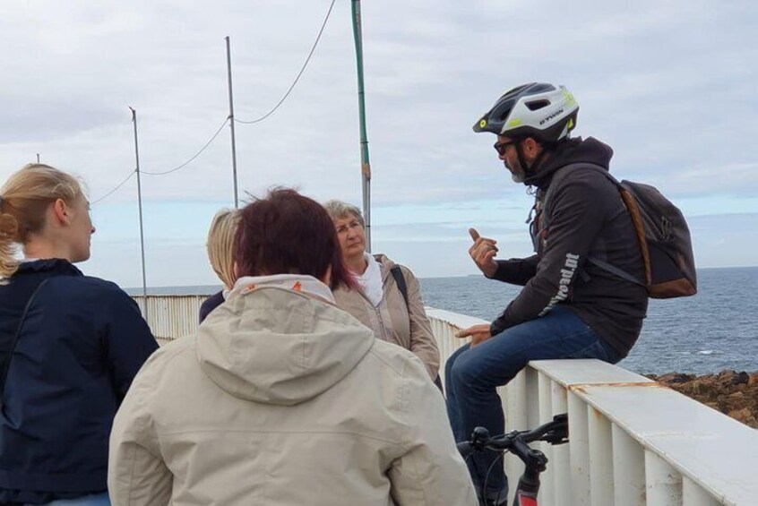 A group of people sitting on a railing by the sea, engaged in conversation. 5
