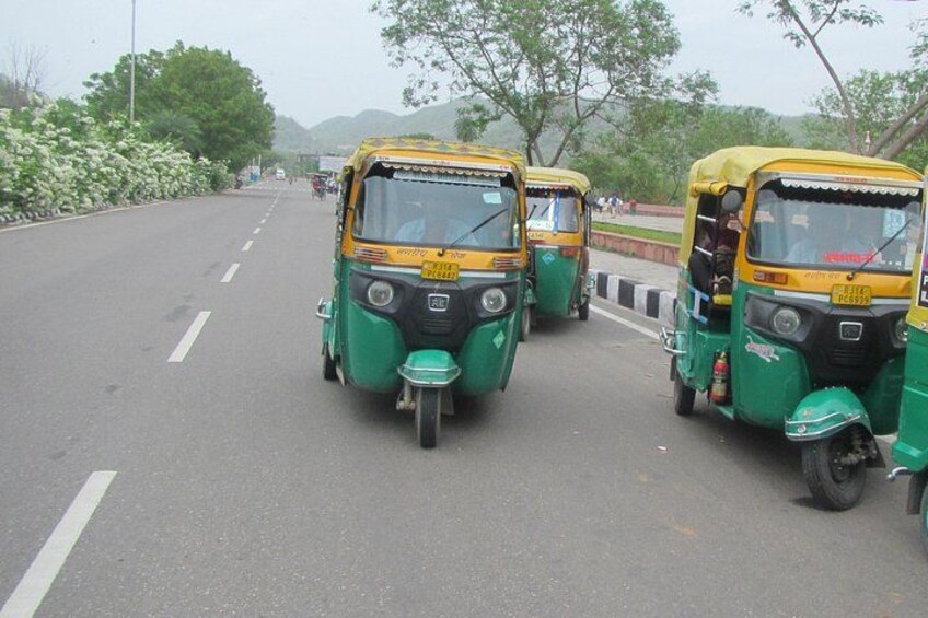 A row of green and yellow auto-rickshaws parked on a road with trees and hills in the background. 5