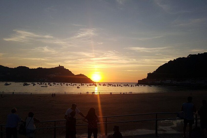A sunset over a beach with people walking along the shore and boats in the water. 1