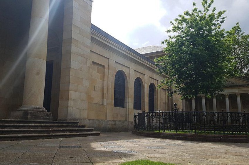 A classical building with columns and arched windows, a paved courtyard, and a tree. 2