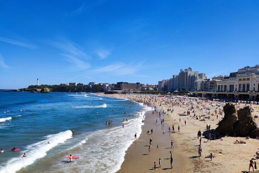 Ein belebter Strand mit badenden und spazierenden Personen, ein Leuchtturm im Hintergrund und Gebäude entlang der Küste. 1