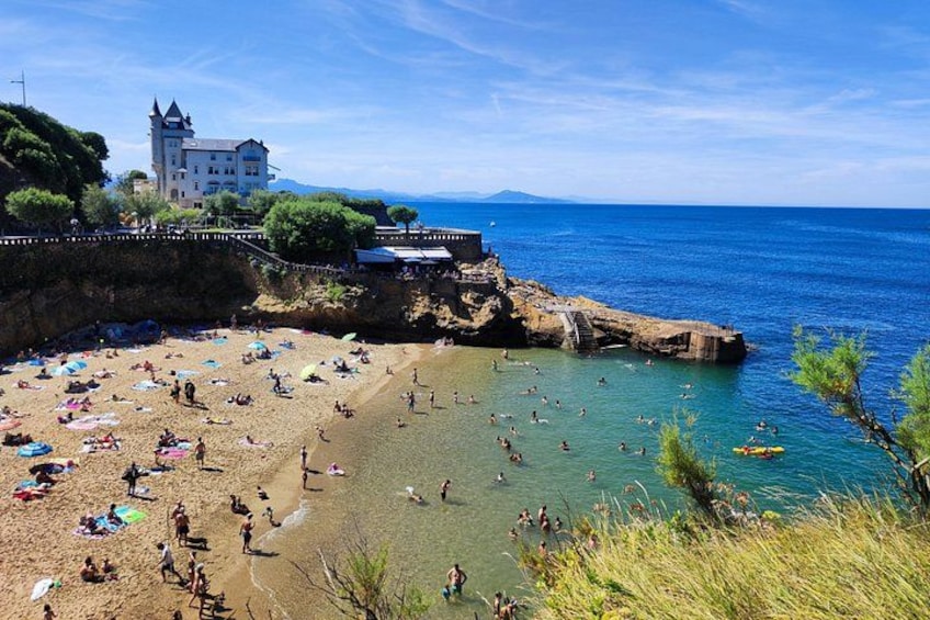 Ein Strand mit badenden und sonnenbäudelnden Menschen, einem Schloss und einem wolkenlosen, blauen Himmel. 5