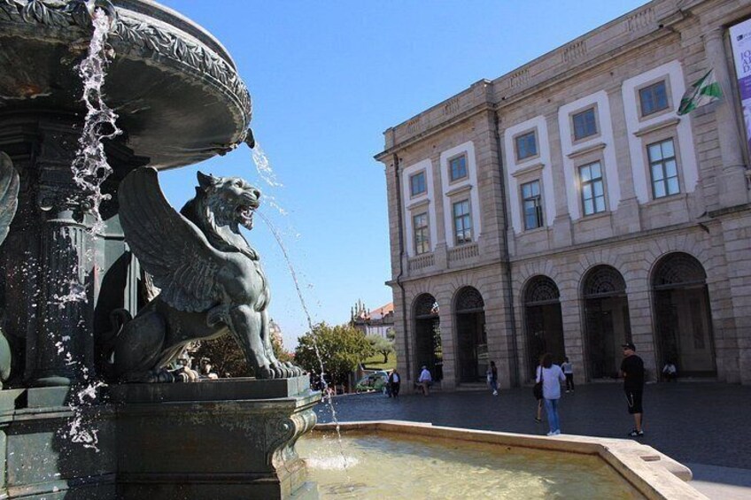 Une fontaine ornée d’une statue de lion, un bâtiment aux fenêtres en arc de cercle et des personnes se trouvant sur une place. 4