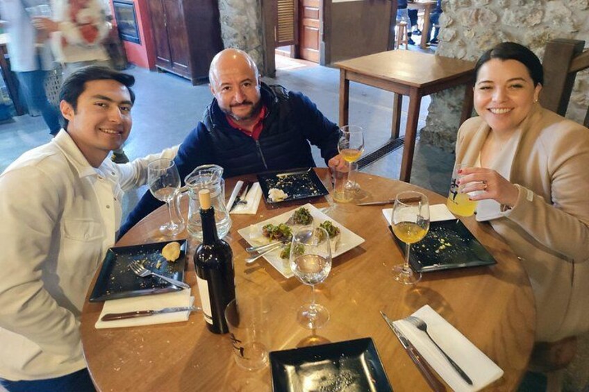 Three people dining at a round table with plates of food, wine glasses, and a bottle of wine. 2
