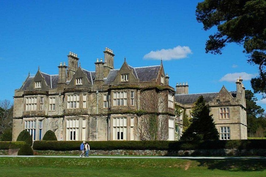 A large, historic stone building with multiple chimneys and windows, surrounded by well-maintained lawns and trees. 1