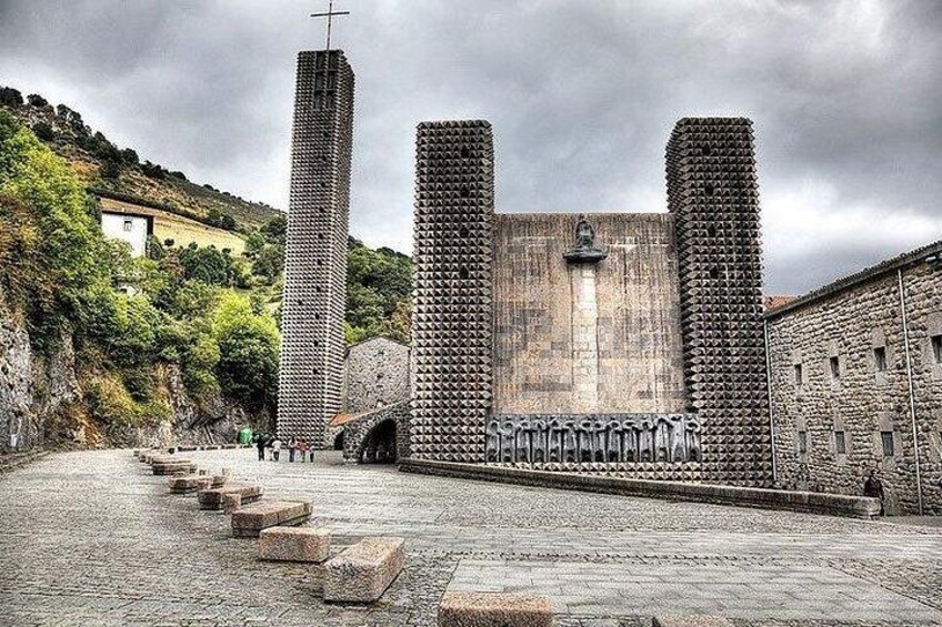Ein steiniger Weg führt zu einem historischen Gebäude mit einem Kreuz auf dem Dach, umgeben von Grünpflanzen und einem bewölkten Himmel. 1