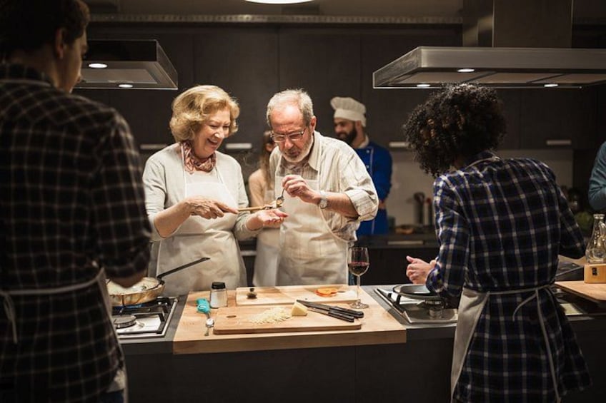 Un gruppo di persone in cucina, alcune con grembiuli, impegnate in attività di cucina o preparazione del cibo. 3