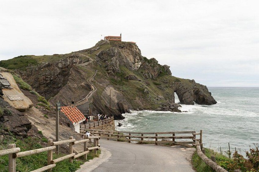 A coastal path leading to a cliff with a building on top, a wooden fence, and a rocky shoreline. 1