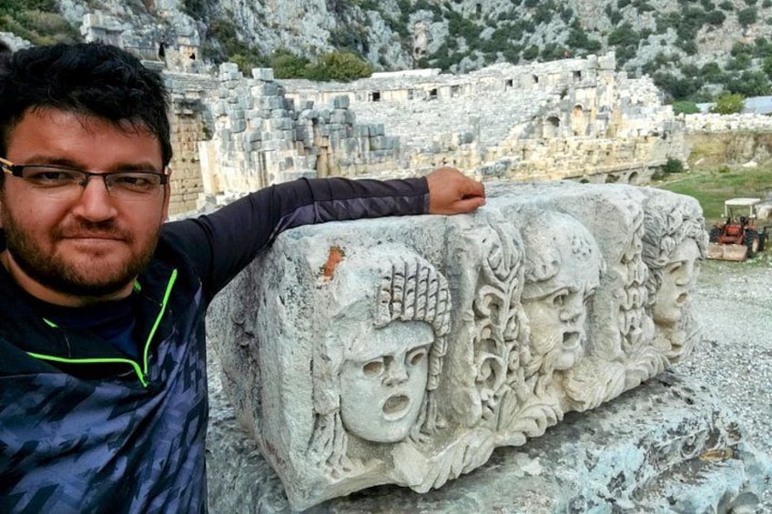 A man leaning on a stone block with carved faces, with ancient ruins in the background. 1