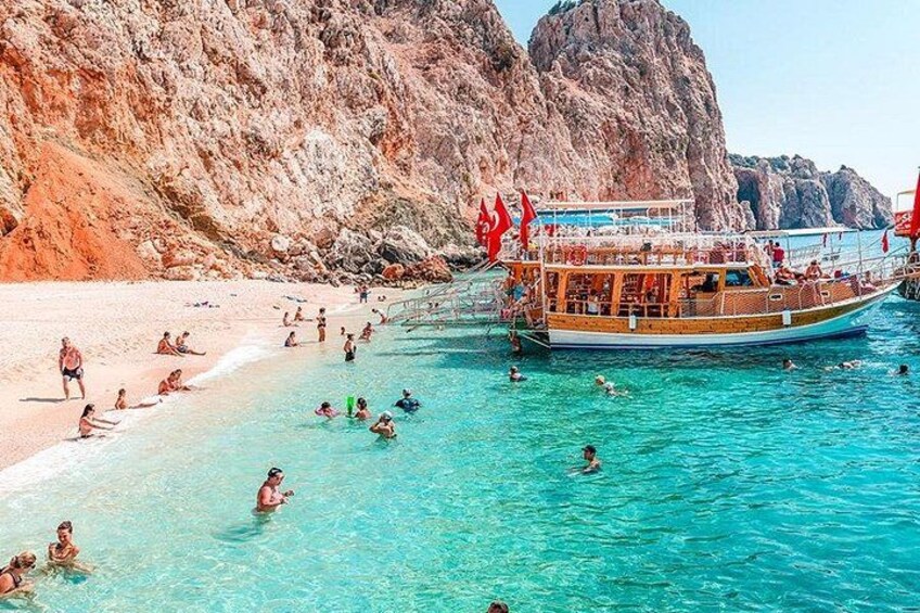 Een strand met helder turquoise water, een boot met rode vlaggen, en rotsachtige kliffen op de achtergrond. 5