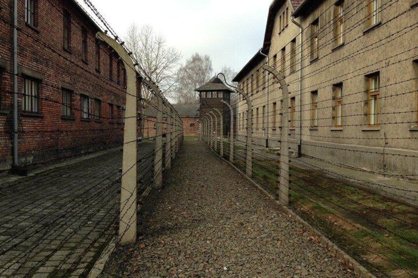 A long corridor with barbed wire fencing on both sides, leading to a building with a tower-like structure in the distance. 3